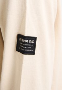 Materialul gri bej al unui hanorac cu un model texturat. Etichetă neagră cusută pe material care afișează "PETROL IND" și alte detalii.