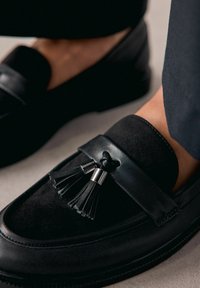 Sorte loafers lavet af læder og ruskind, med en frynsedetalje og sølvfarvet hardware. Glat overflade med rund tå og flad sål.