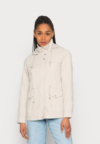 Chaqueta con capucha de color beige claro, con cordones, cierre de cremallera y dos bolsillos en el pecho con botón. Textura suave, mangas largas y ajuste casual.