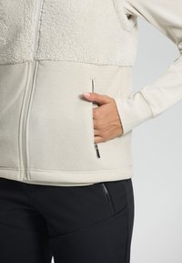 Ljusbeige väst med mjukt, fluffigt övre tyg och slät underdel. Har en sidoficka med dragkedja. Kombinerad med svarta byxor.