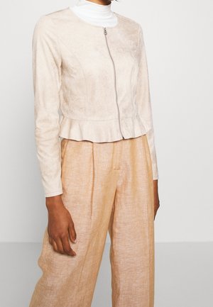 Femme portant une veste zippée beige clair avec un bas à volant, sur un col roulé blanc et un pantalon ample beige, sur un fond uni.