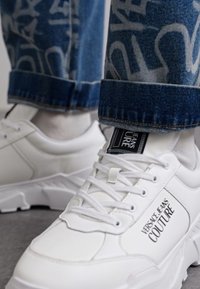 Sapatilhas brancas chunky feitas de couro liso com uma solado texturizado. Apresentam um logo preto "Versace Jeans Couture" na lateral.