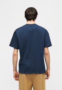 Άνδρας που φοράει μπλε σκούρο κοντομάνικο μπλουζάκι και μπεζ παντελόνι, στέκεται με την πλάτη στην κάμερα μπροστά σε απλό φωτεινό φόντο.