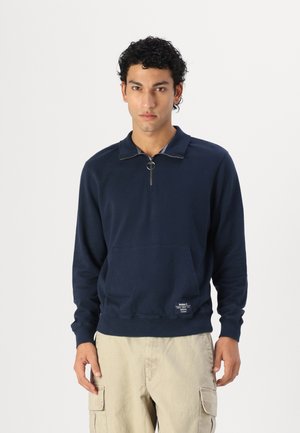 Pullover blu marina con zip a un quarto, realizzato in un tessuto morbido, dotato di una tasca frontale e di una piccola etichetta sull'orlo. Presenta un design semplice e maniche lunghe.