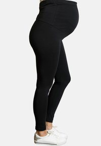 Leggings de maternidad negros hechos de tela elástica con una cintura alta. Combinados con zapatillas blancas, mostrando un diseño ajustado y estilizado.