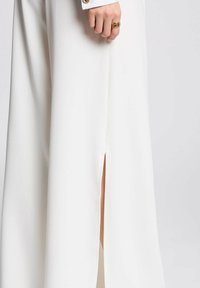 Pantaloni bianchi a gamba ampia con spacchi laterali. Realizzati in tessuto morbido e liscio. Presentano una manica con risvolto e dettaglio di bottone decorativo.