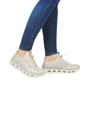 Beige athletic shoes with mesh and synthetic overlays, featuring a textured sole and lace-up design, worn with dark blue fitted jeans.