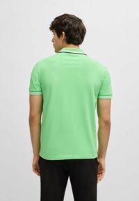 Grön pikétröja i texturerat tyg, med klassisk krage och svarta accentstripes på ärmarna. Sedd från baksidan.