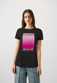 Zwarte t-shirt met een gradiënt roze en witte rechthoekige logo met "GUESS" in vetgedrukte zwarte letters en een decoratieve zilveren schrift eronder.