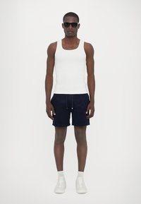 Débardeur blanc, shorts navy avec cordon de serrage, baskets blanches et lunettes de soleil noires. La tenue présente un design ajusté et un style décontracté.