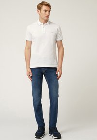 Polo bianco con colletto e maniche corte, abbinato a jeans blu e scarpe da ginnastica nere, su uno sfondo neutro.