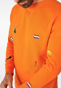 Oranžinė megztinis su įvairiais siuvinėjimais: vėjo malūnas, Olandijos vėliava, futbolo kamuolys, gėlė, olandiškas sūris ir batelis. Minkšta tekstūra.