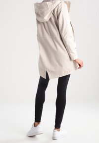 Vaaleanbeige sadetakki hupulla, jossa on istuva vyötärö, sivuhalkiot ja sileä pinta, yhdistetty mustiin legginsseihin ja valkoisiin lenkkareihin.