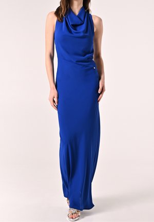 Vrouw draagt een mouwloze, koningsblauwe avondjurk tot op de grond met een gedrapeerde halslijn en zilveren open-teenhakken, staand tegen een effen achtergrond.