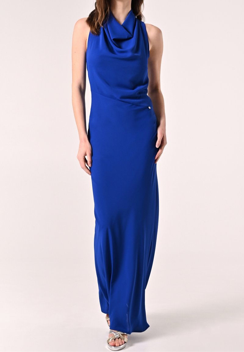 Femme portant une robe longue sans manches bleu roi avec un décolleté drapé et des talons argentés ouverts, debout devant un fond uni.