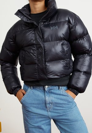 Personne portant une doudoune noire brillante avec un logo sur la poitrine et un jean bleu clair, mains dans les poches, sur fond uni.