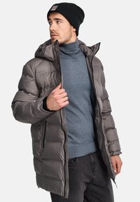 Chaqueta gris acolchada con capucha, que presenta un cierre de cremallera y puños elásticos, llevada sobre un cuello de tortuga azul y jeans negros.