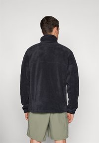 Veste en polaire bleu marine foncé avec un col montant et des manches longues, présentant une surface texturée. Portée avec un short vert clair.