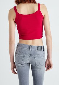 Une personne portant un crop top sans manches rouge et un jean ajusté gris clair, vue de dos sur un fond blanc uni.