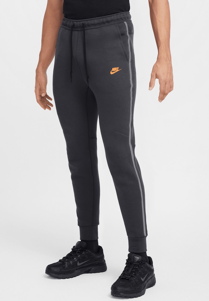 Pelēkas sporta bikses ar elastīgu jostu, sānos ar svītrām un oranžu Nike logo. Valkātas ar melniem sporta apaviem.