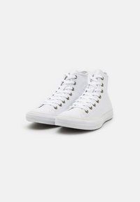 Białe trampki typu high-top z płótna, z metalowymi przelotkami, płaskimi sznurówkami, teksturowaną gumową podeszwą oraz kontrastującą białą i beżową linią wzdłuż podstawy.