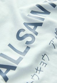 Ljusblå bomullströja med stor mörkblå text som säger "ALL SAINTS" och mindre text i en annan stil nedanför. Slät textur.