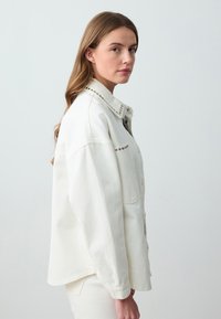 Chemise en denim blanche avec une coupe décontractée, ornée de clous décoratifs le long du col et sur le devant. Manches longues, fermeture à boutons et tissu texturé.