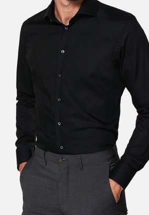 Mężczyzna w dopasowanej czarnej koszuli na guziki z długim rękawem, wpuszczonej w szare spodnie wizytowe, z rękami w kieszeniach.