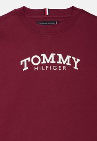 Burgundi puuvillane dressipluus, millel on ümmargune kaelus, valge "TOMMY HILFIGER" logo rinna keskosas ja triibuline aksent kaeluse serval.