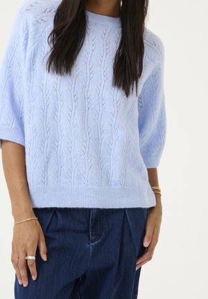 Pull en tricot bleu clair à manches courtes avec un motif de feuilles texturé, associé à un jean plissé taille haute bleu foncé.