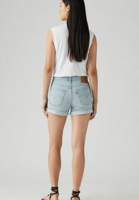 Shorts de mezclilla azul claro con puños enrollados, con dos bolsillos traseros y una etiqueta de cuero marrón en la cintura. Se lleva una blusa blanca sin mangas.