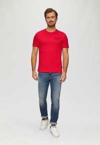 Rode t-shirt met korte mouwen met de tekst "FISH N' CHIPS", gecombineerd met lichtblauwe jeans en witte sneakers. Gladde stof; casual pasvorm.