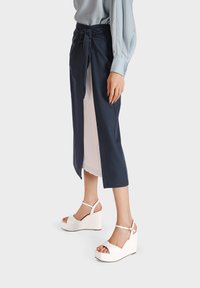 Gonna avvolgente blu navy con sottogonna plissettata bianca, legata in vita. Sandali con zeppa bianchi con cinturini alla caviglia e suole larghe.