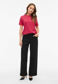 Dopasowany różowy T-shirt z krótkim rękawem w połączeniu z czarnymi, szerokimi spodniami z wysokim stanem oraz czarnymi, szpiczastymi obcasami, stojąc na jednolitym tle.