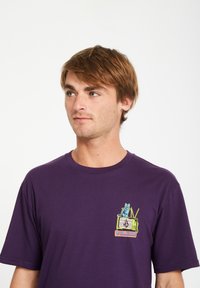 Lila Baumwoll-T-Shirt mit einem Frontgrafik eines blauen Katzen auf einem vintage Fernseher, mit dem Wort "VOLCOM" in buntem Text.