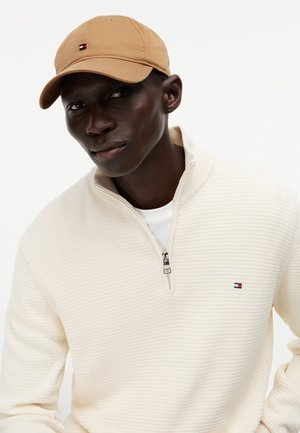 Uomo con cappellino beige e maglione crema a mezza zip con tessitura, con piccolo logo rosso, bianco e blu sul petto, su sfondo bianco.