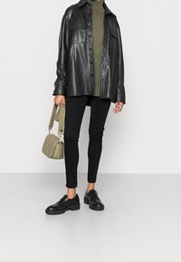Giacca camicia di pelle nera sopra un dolcevita verde, abbinata a jeans skinny neri e mocassini neri voluminosi. Tiene una borsa a tracolla trapuntata verde oliva.