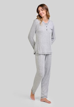 Mujer de pie descalza, vestida con un conjunto de pijama de punto gris claro de manga larga con botones y un bolsillo en el pecho, sonriendo y mirando hacia abajo.