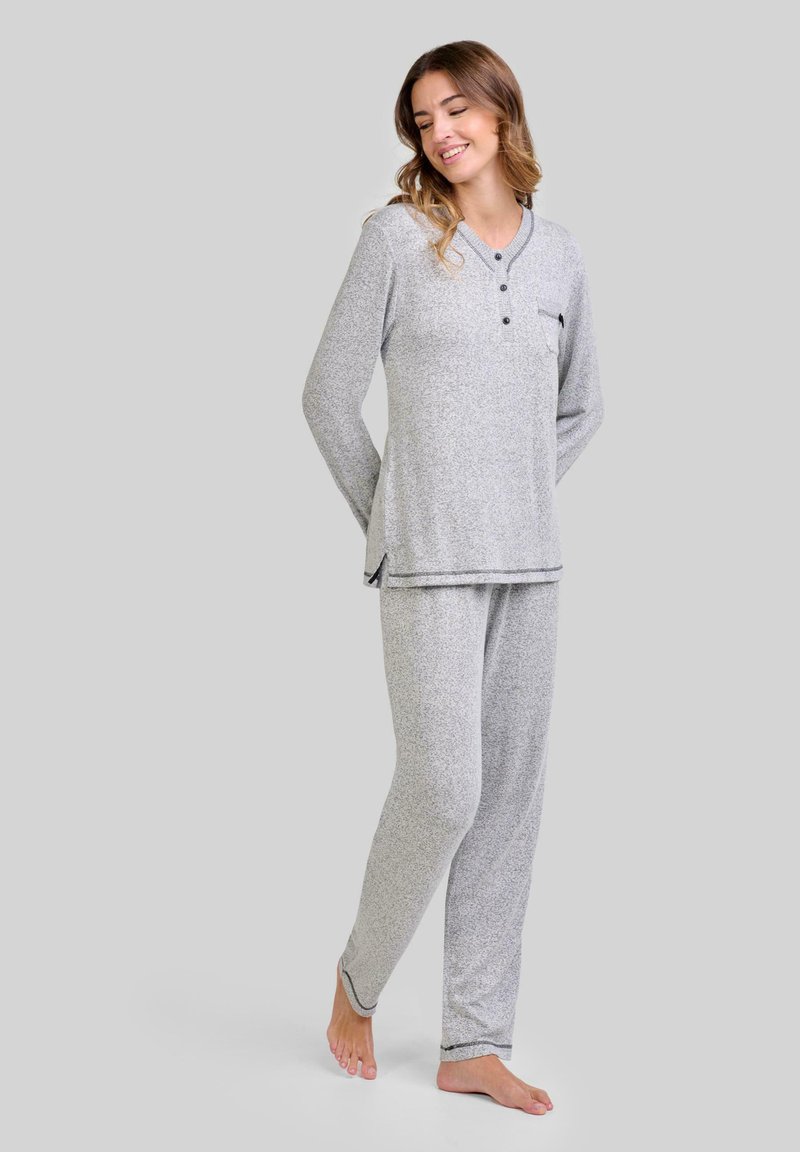Mujer de pie descalza, vestida con un conjunto de pijama de punto gris claro de manga larga con botones y un bolsillo en el pecho, sonriendo y mirando hacia abajo.