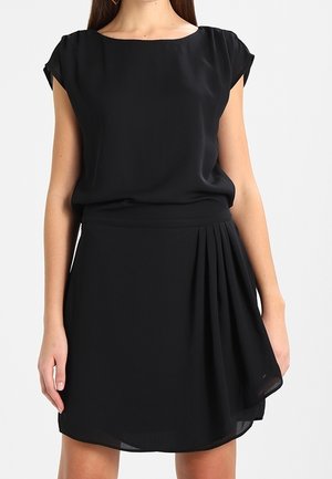 Femme portant une robe noire courte avec des manches courtes et un détail plissé sur un côté de la jupe.