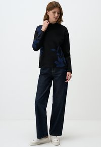 Zwarte lange mouwen top met donkerblauw bloemenpatroon, hoge halslijn, gecombineerd met losse donkerdenim jeans en witte sneakers, staande pose.