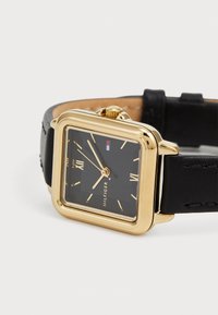 Montre carrée dorée avec un cadran noir, des chiffres romains et des aiguilles dorées. Dispose d'un bracelet en cuir noir et d'un logo de la marque sur le cadran.