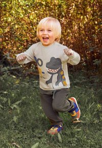 Kind in een lichtgrijze sweater met een dinosaurusafbeelding, gecombineerd met gestructureerde donkergroene broek en meerkleurige sportschoenen, die buiten rent.