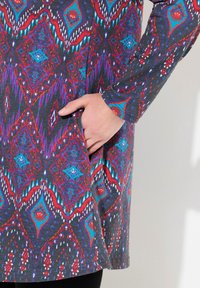 Langdraagjurk met een meerkleurig geometrisch patroon in blauw, rood en paars op een donkergrijze achtergrond, met een zijzak.