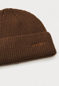 Căciulă tricotată ribbed, de culoare maro, cu margine pliată, având un logo brodat subtil într-o nuanță mai deschisă, confecționată dintr-un amestec moale de lână texturată.