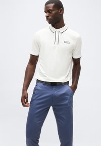 Άνδρας με λευκό κοντομάνικο πόλο με μαύρο περιθώριο και μπλε παντελόνι, στέκεται με το ένα χέρι στην τσέπη, κοιτώντας αριστερά του, μπροστά σε μονόχρωμο φόντο.