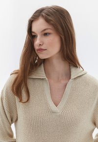 Beige stickad tröja med V-ringning och krage, med ett mönster och löst snitt. Mjuk material, medellånga ärmar, avslappnad design.