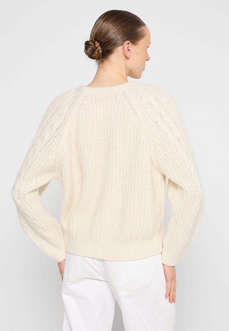 Frau mit geflochtenem Dutt, die einen cremfarbenen Strickpullover mit Kabelmustern an den Ärmeln und weiße Hose trägt, von hinten gegen einen schlichten Hintergrund blickend.