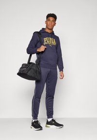 Jūrinė bliuzono su "PUMA sporto klubo" logotipu, derančios siaurėjančios treniruotės kelnės su šoninėmis juostel�ėmis ir juodas sportinis krepšys. Avalynė: juodi sportbačiai.