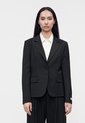Vrouw met lang donker haar draagt een zwarte blazer over een witte blouse met knopen, staand tegen een effen lichte achtergrond.
